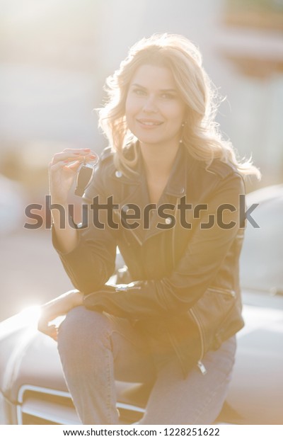 Photo of young blonde woman with keys sitting on hood\
of black car