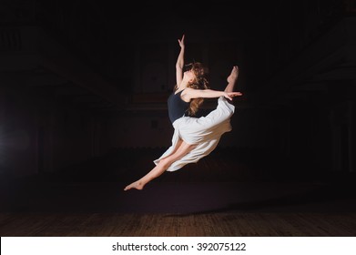 Photo Of Young Brunette Dancer Girl, Ballerina In White Skirt In Split Jump On Stage In Theater With Spotlight. Barefoot Dancer.