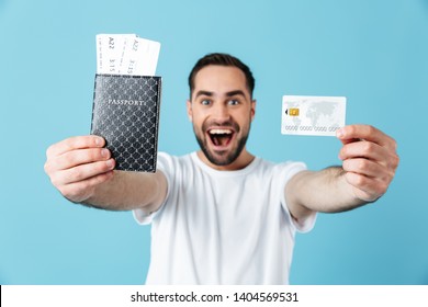 Photo Of Young Brunette Man Wearing Basic T-shirt Smiling While Holding Travel Tickets With Passport And Credit Card Isolated Over Blue Background