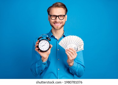 Photo of young business man happy positive smile hold clock time cash rich dollars isolated over blue color background