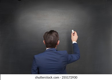 Photo Of Young Businessman On Blank Chalkboard Background. Man Standing With His Back To The Camera And Writing With Chalk