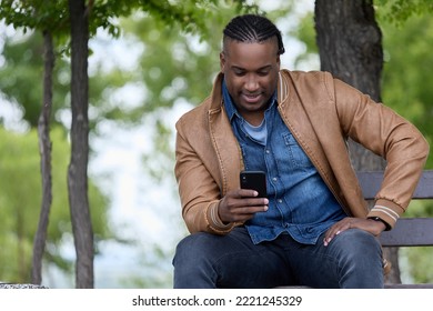 Photo of young businessman in a stylish brown bomber strolling through a green park with a smartphone on a sunny day. Beautiful African businessman sits on the street in a park and uses his phone - Shutterstock ID 2221245329