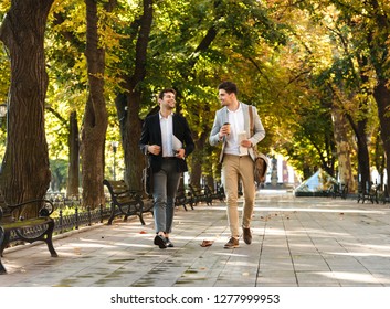 Photo of young businessmen in suits walking outdoor through green park with takeaway coffee and laptop during sunny day