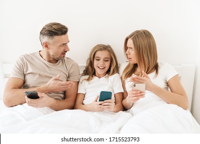 Photo Of Young Caucasian Family Expressing Shock And Using Cellphones While Sitting In White Bad After Sleep