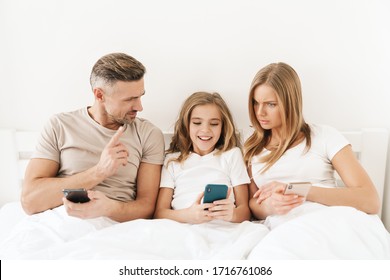 Photo Of Young Caucasian Family Expressing Shock And Using Cellphones While Sitting In White Bad After Sleep