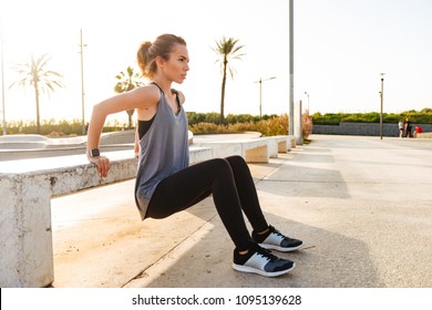 Photo of young caucasian girl in sportswear squatting, and doing workout using concrete bench in sport park at sunrise