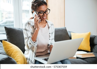 Photo Of Young Cheerful African Woman Wearing Glasses Sitting In Library Looking At Laptop And Talking By Phone.