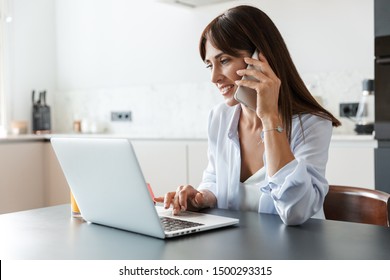 Photo of a young cheerful happy woman indoors at home at the kitchen using laptop computer talking by mobile phone.