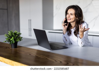 Photo of a young cheerful happy woman indoors at home at the kitchen using laptop computer talking by mobile phone. - Shutterstock ID 2232585687