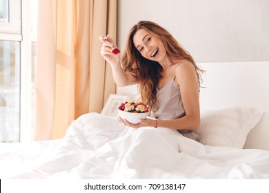 Photo Of Young Cheerful Pretty Lady Sitting On Bed Indoors. Looking Camera Eating Fruit Salad.