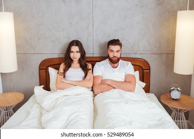 Photo Of Young Confused Loving Couple Lies In Bed With Arms Crossed. Looking At Camera.