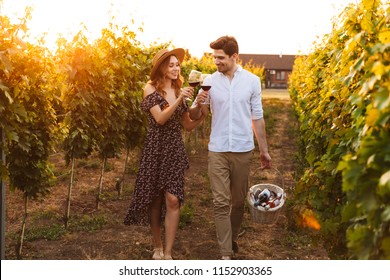 Photo Of Young Cute Happy Loving Couple Outdoors Drinking Wine Holding Basket With Bottles.