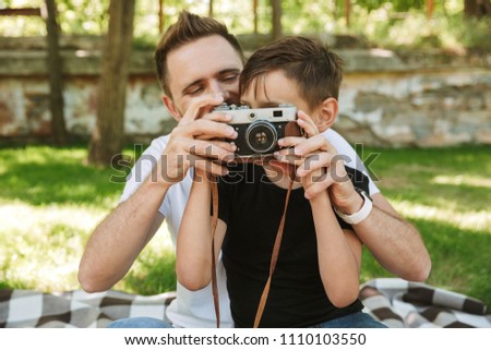 Photo of young father sitting with his little son outdoors in park nature holding camera photographing.
