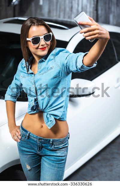 Photo of young female car repair worker. Glamour\
sexy brunette wearing jeans. Girl with sunglasses making selfie\
photo on mobile phone near\
car