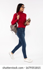 Photo Of Young Female Student Walking With A Backpack And Smiling, Holding Books In Her Hands, Wearing Casual Denim Pants, Red Sweater And Reading Glasses, Isolated On White Background