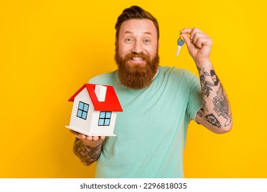 Photo of young funny excited happy man wear green t-shirt hold his new property keys miniature house for sale isolated on yellow color background