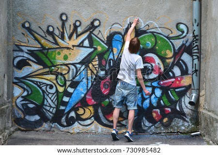 Photo of a young guy in denim shorts and a white shirt. The guy draws on the graffiti wall a drawing with aerosol paints of various colors. The concept of hooliganism and damage to property