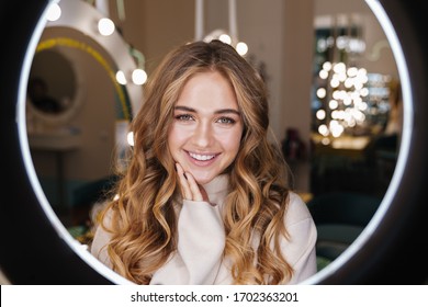 Photo Of A Young Happy Cute Blonde Girl Indoors In Beauty Salon Looking At Camera Through Ring Light Lamp.