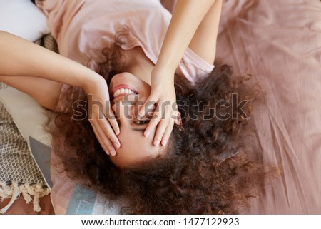 Photo of young happy dark skinned lady lying on the bed and covers face with hands, enjoy the sunny day at home and smiling, spends she's free day at home.