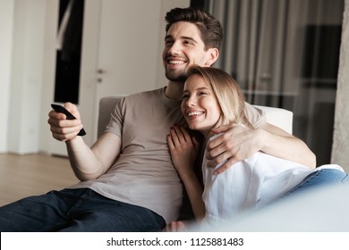 Photo Of Young Happy Loving Couple In Home Indoors On Sofa Watch TV Holding Remote Control.