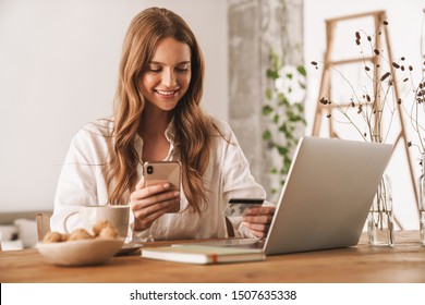 Photo Of Young Happy Redhead Business Woman Sit Indoors In Office Using Laptop Computer And Mobile Phone Holding Credit Card.