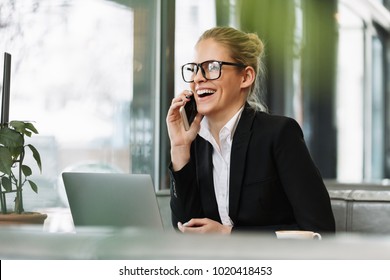 Photo of young happy smiling blonde business woman sitting indoors in cafe using laptop computer talking by mobile phone. Looking aside.
