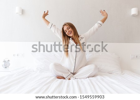 Photo of young happy woman in pajama stretching her arms and smiling while sitting on bed after sleep or nap