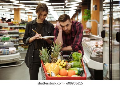 Photo of young loving couple in supermarket with shopping trolley choosing products. Tired man looking at camera. - Powered by Shutterstock