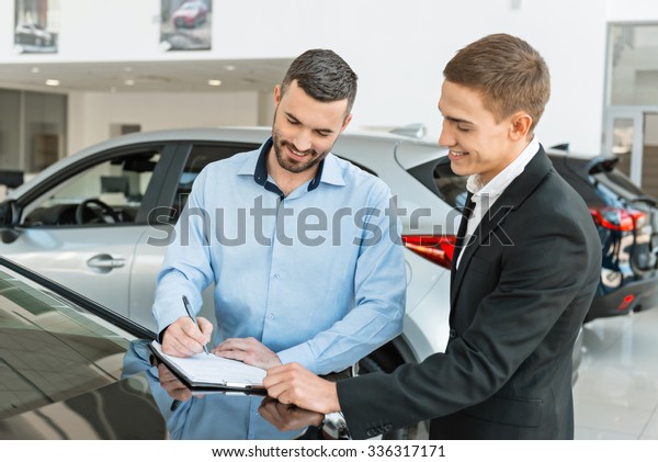 Photo of young male\
consultant and buyer signing contract for new car in auto show.\
Concept for car rental