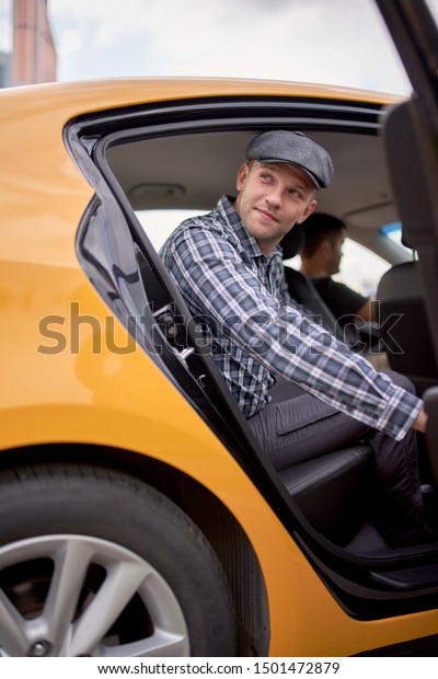 Photo of young man in cap and plaid\
shirt looking to side sitting in back seat in yellow\
taxi