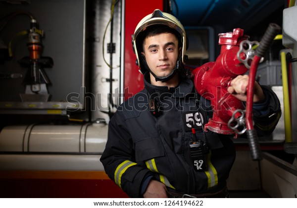 Photo of young man firefighter\
with fire cock on shoulder against background of fire\
truck