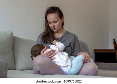 Photo of a young mother breastfeeding her kid sitting against light living room at home. Young mother holding her baby child. Mom nursing baby. Young mother holding and nursing her newborn child.