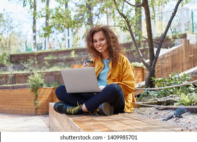 Photo of young nice dark skinned curly girl, sits in the park with legs crossed and drinks coffee, with a laptop on lap, looks at the camera and smiling.