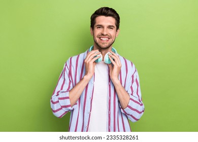 Photo of young optimistic man wearing stylish striped shirt touching his wireless earphones itunes isolated on green color background