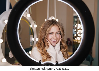 Photo Of A Young Positive Cute Cheery Blonde Girl Indoors In Beauty Salon Looking At Camera Through Ring Light Lamp.