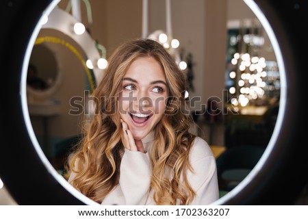 Photo of a young positive cute cheery blonde girl indoors in beauty salon posing at camera through ring light lamp.