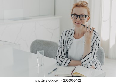 Photo of young red-haired smiling businesswoman using voice assistant app on smartphone, sitting at table at home and planing her day in morning, making notes in notebook and drinking mineral water - Powered by Shutterstock