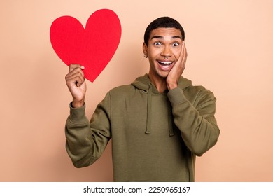 Photo of young romantic funny guy excited positive touch cheek hold red paper love symbol like his partner isolated on beige color background