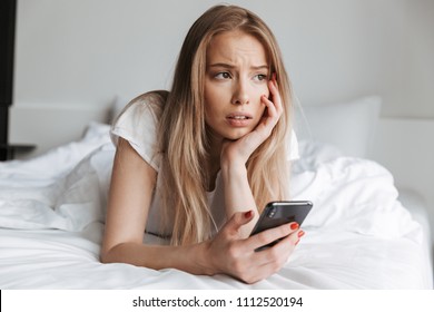 Photo Of Young Sad Woman Lies On Bed Indoors At Home Using Mobile Phone. Looking Aside.