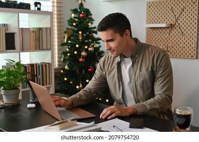 Photo Of A Young Smart Man Using A Computer Laptop At The Cluttered Desk Over The Christmas Tree And Bookshelf As A Background.