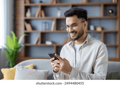 Photo of a young smiling Indian man sitting at home on the sofa and using a mobile phone in his hands. - Powered by Shutterstock