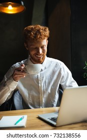 Photo Of Young Smiling Readhear Bearded Man Holding Coffe Cup, Looking At Laptop Screen While Working At Cafeteria