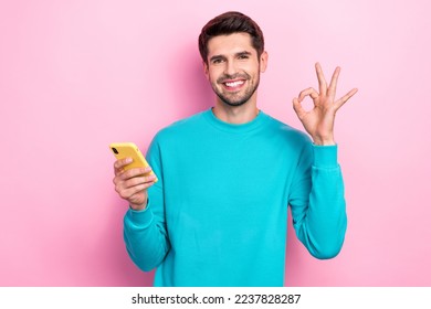 Photo of young smiling satisfied brunet hair businessman wear blue sweater show okey sign smartphone recommend shop isolated on pink color background - Powered by Shutterstock