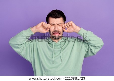 Photo of young unhappy funny cute man bearded brunet crying loud grimace wipe tears dont like bad news childish isolated on violet color background