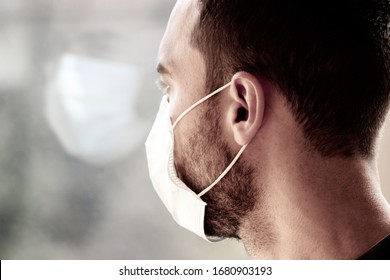 Photo Of A Young White Man With Medical Face Mask Looking Out Of The Window During Coronavirus Quarantine. Reflection Of His Face In The Window. Coronavirus, COVID-19 Outbreak. Doctor, Nurse Concept.