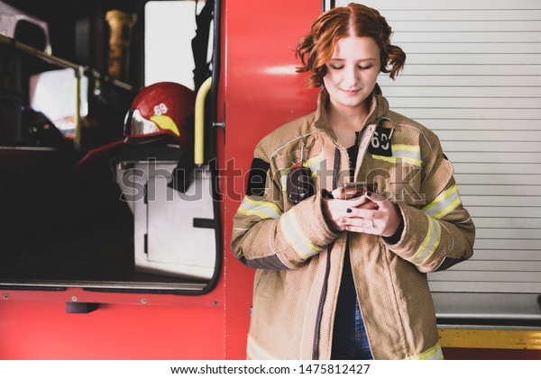 Photo of young woman\
firefighter with phone in her hands against background of fire\
engine