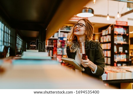 Photo of young woman with glasses returning book to the book shelf. Young woman is looking for book to buy at bookstore.