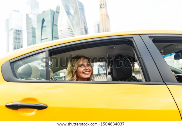 Photo of young woman sitting in\
back seat of yellow taxi in summer on modern city\
background.