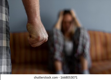 Photo Of Young Woman Sitting On Sofa At Home,focus Is On Man's Fist In The Foreground Of The Image.Home Violence Concept.Frightened Woman And Men's Fist.Woman Is Victim Of Domestic Violence And Abuse.