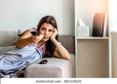 Photo of a young woman sitting on a sofa, feeling depressed and changing channels on a TV remote. Young brunette woman relaxing at home, curled up in a blanket, holding the remote, watching television